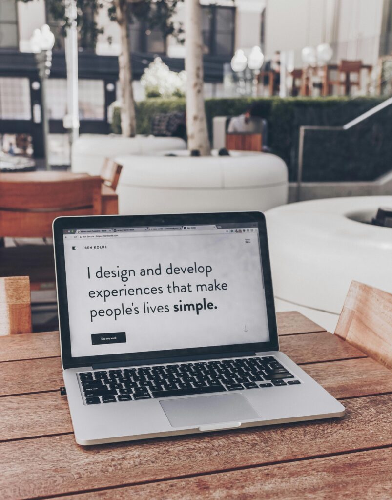 A laptop sitting on a wooden outdoor table displaying the text: 'I design and develop experiences that make people's lives simple.' emphasizing clean, user-centered design.
