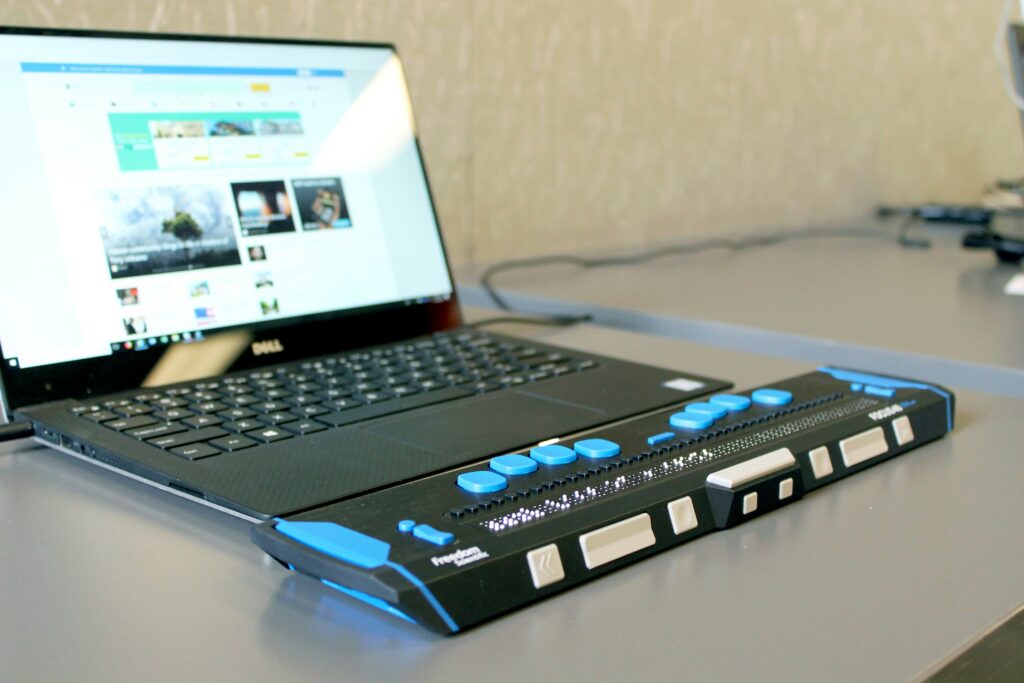 A high-tech refreshable Braille display device with blue and white tactile buttons connected to a laptop, demonstrating assistive technology for blind or visually impaired users.