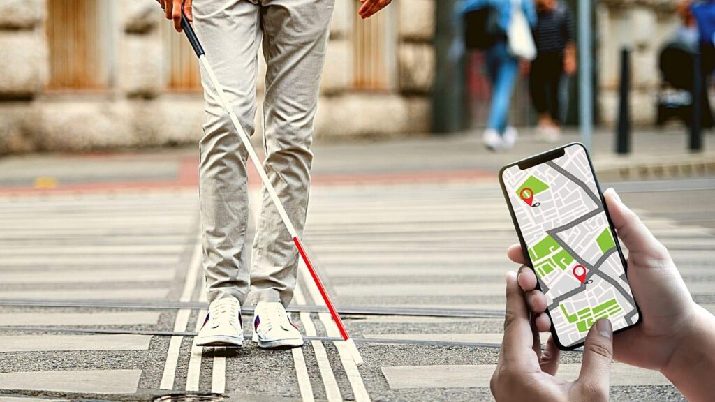 Low-angle shot of a person using a white cane to navigate a tactile paving crosswalk, while a hand in the foreground holds a smartphone displaying a GPS navigation map with location pins.